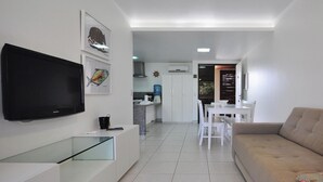 Uma sala de estar moderna com televisão de tela plana, mesa de centro de vidro, sofá e um espaço de refeições com mesa e cadeiras brancas.