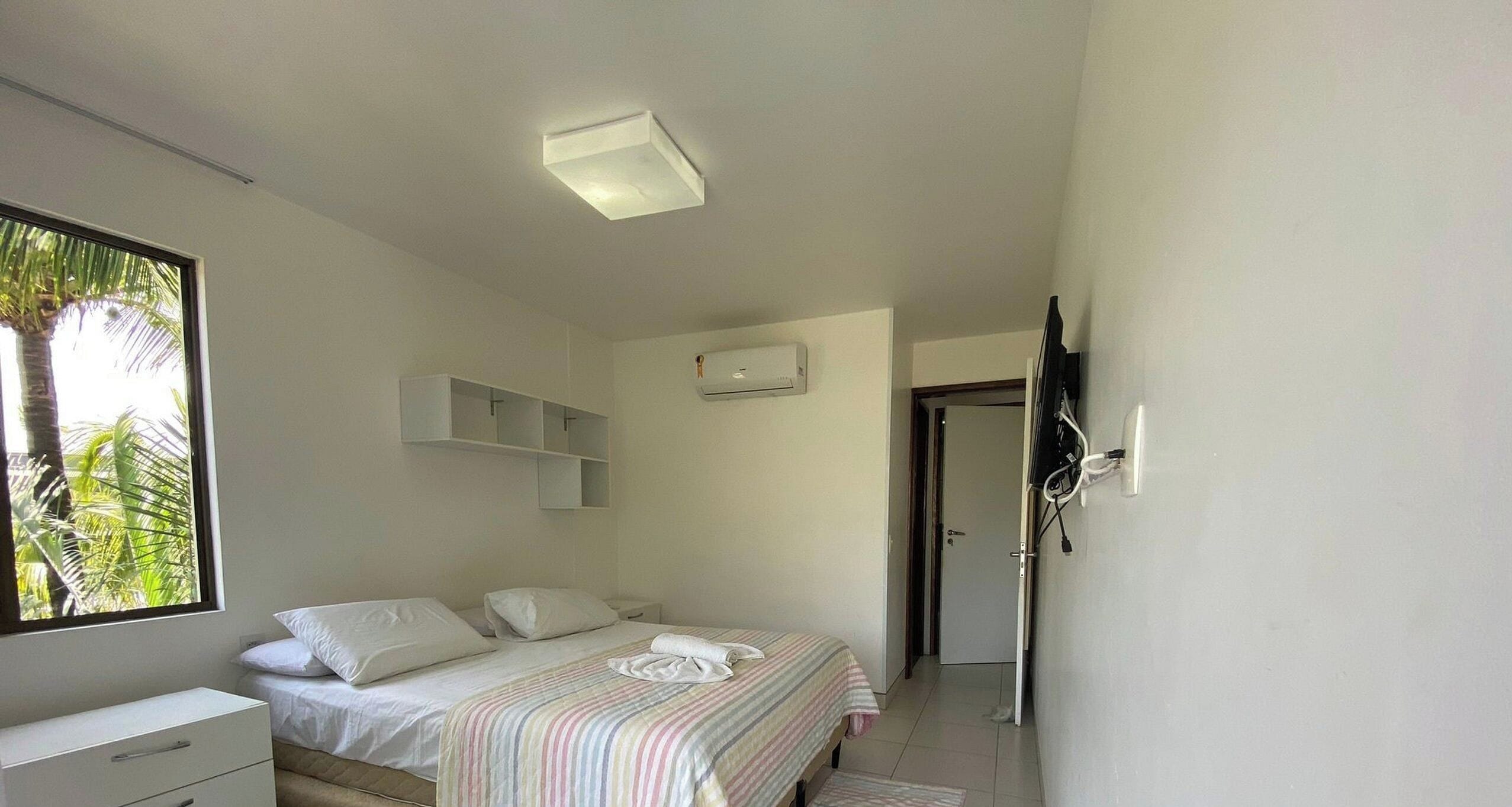 A bedroom with a bed, a bedside table, a wardrobe, and a window with a view of palm trees.