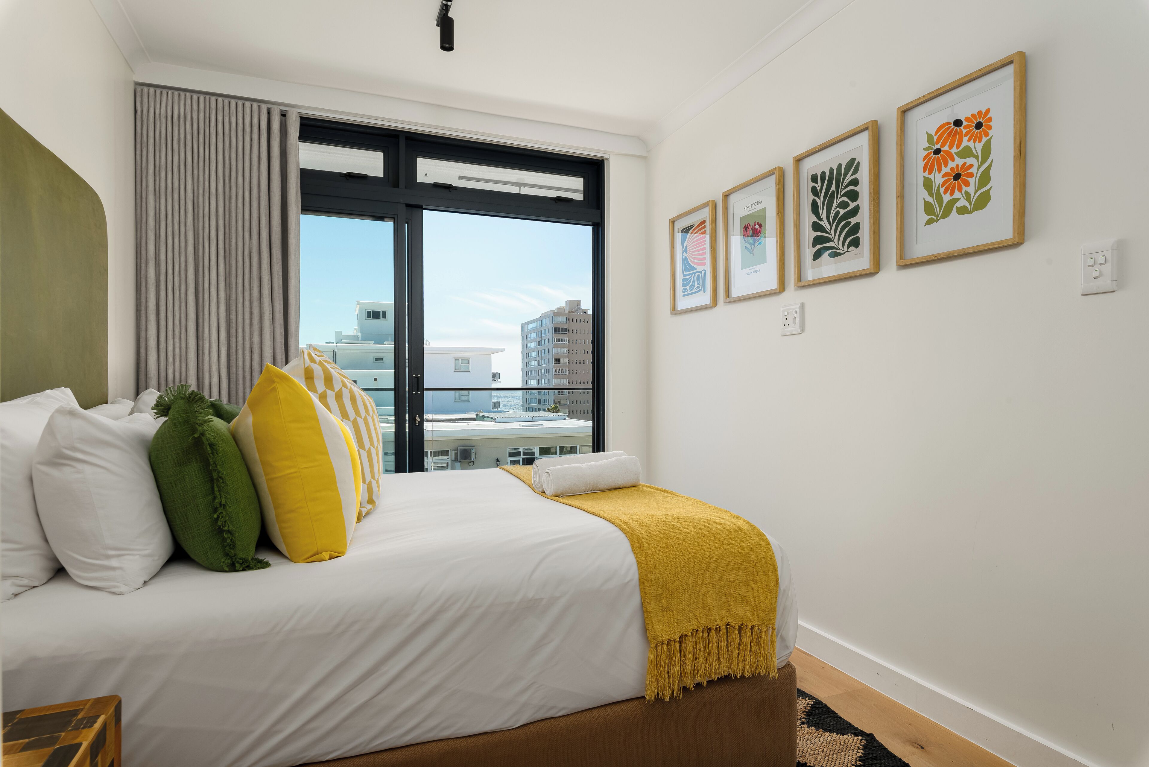 A hotel room with a large bed, a view of buildings through a window, and three framed artworks on the wall.