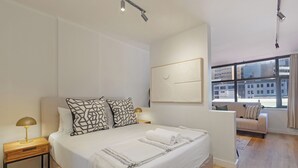 Et moderne soveværelse med en stor seng, sengeborde, en sofa og udsigt over byen gennem store vinduer.
