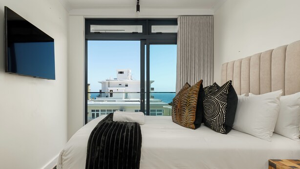 A hotel room with a large bed, a flat-screen TV mounted on the wall, and a view of the sea and buildings through the window. - Stylish Apartment in the Heart of Sea Point (Cape Town)