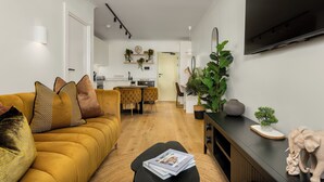 A modern living room with a sofa, a coffee table, and a flat-screen TV. There is a dining area with a table and chairs, and a kitchen in the background. - Stylish Apartment in the Heart of Sea Point (Cape Town)