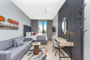 A modern hotel room with a grey sofa, a bed with grey bedding, a wooden desk, and a large window offering a city view. - Sleek Luxury Studio at One Rosebank (Johannesburg)
