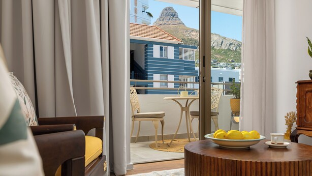 Une pièce avec un balcon, une table en bois sur laquelle se trouve un panier de citrons, et une vue sur un bâtiment avec un toit rouge et une montagne en arrière-plan.