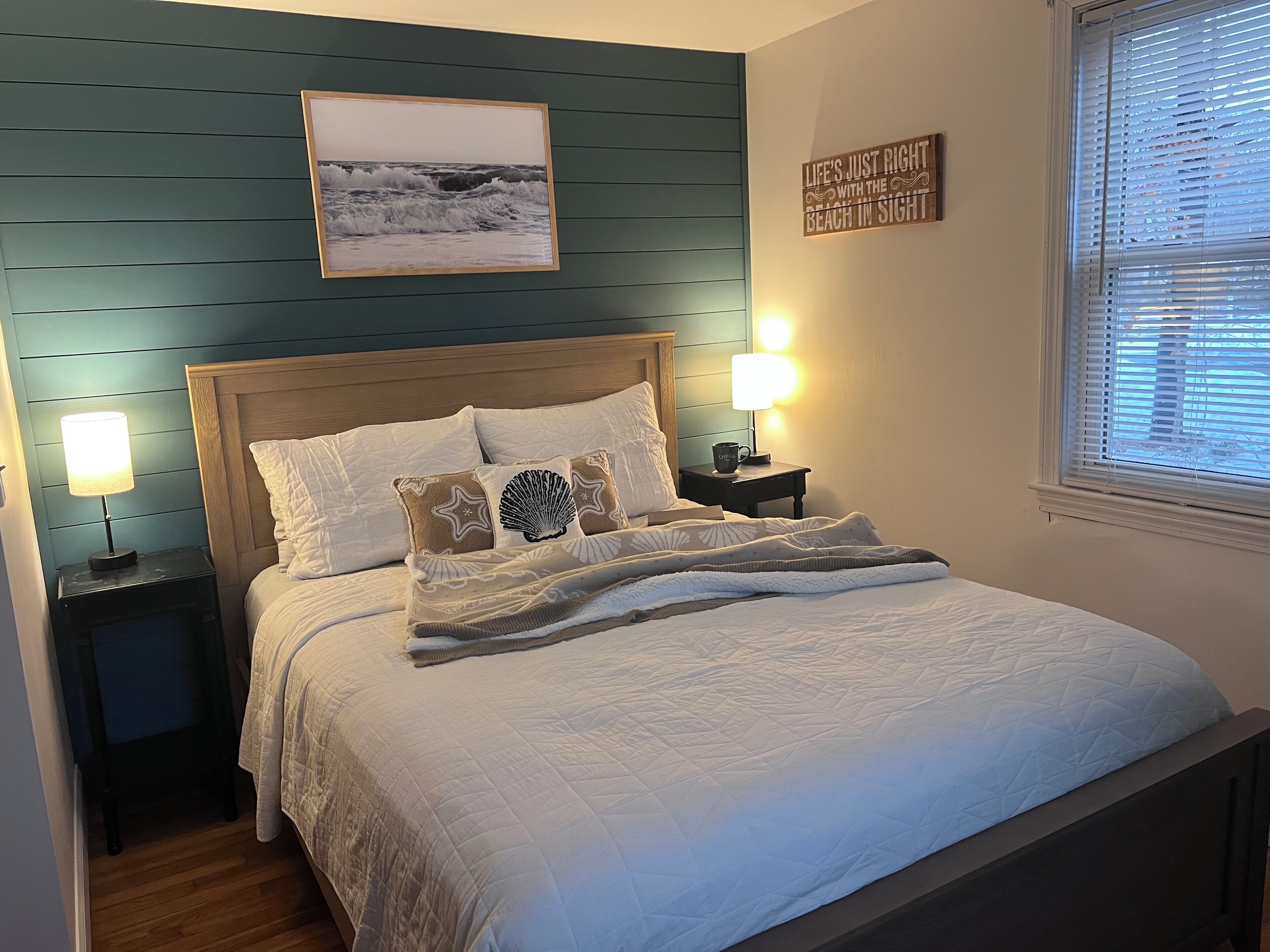 A neatly made bed with white bedding and decorative pillows, a framed picture above the bed, a bedside lamp, and a window with blinds.