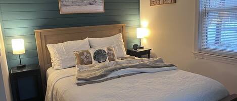 A neatly made bed with white bedding and decorative pillows, a framed picture above the bed, a bedside lamp, and a window with blinds.