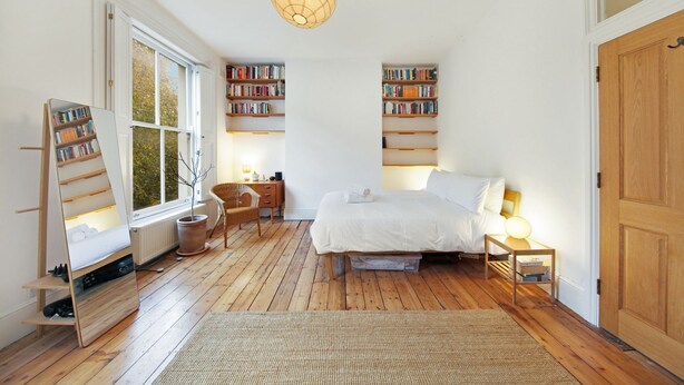 Une chambre avec un grand lit, un plancher en bois, un miroir, une bibliothèque et une fenêtre donnant sur des arbres.