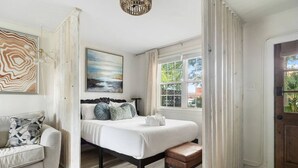 Ein Schlafzimmer mit Bett, Stuhl, Gemälde und einem Fenster mit Vorhängen.