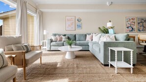 Un salon comprenant un canapé d’angle, une table basse ronde et une petite table blanche. On y trouve deux tables d’appoint avec des lampes, une plante en pot et des œuvres d’art accrochées aux murs.