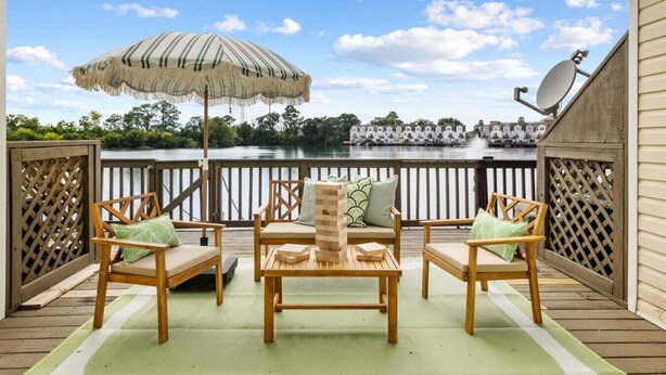 Une terrasse avec des chaises et une table en bois, un parasol à rayures et une vue sur un lac, avec des bâtiments en arrière-plan.