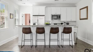 Una cocina moderna con armarios blancos, un refrigerador de acero inoxidable, un microondas y una encimera de granito. Hay cuatro taburetes de bar con estructura metálica y asientos marrones.