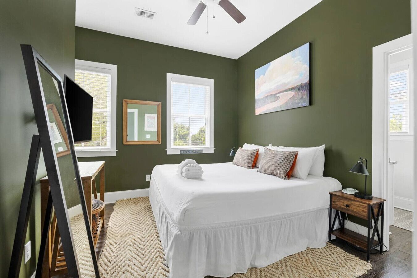 A bedroom with a large bed, two windows, a ceiling fan, and a painting on the wall.
