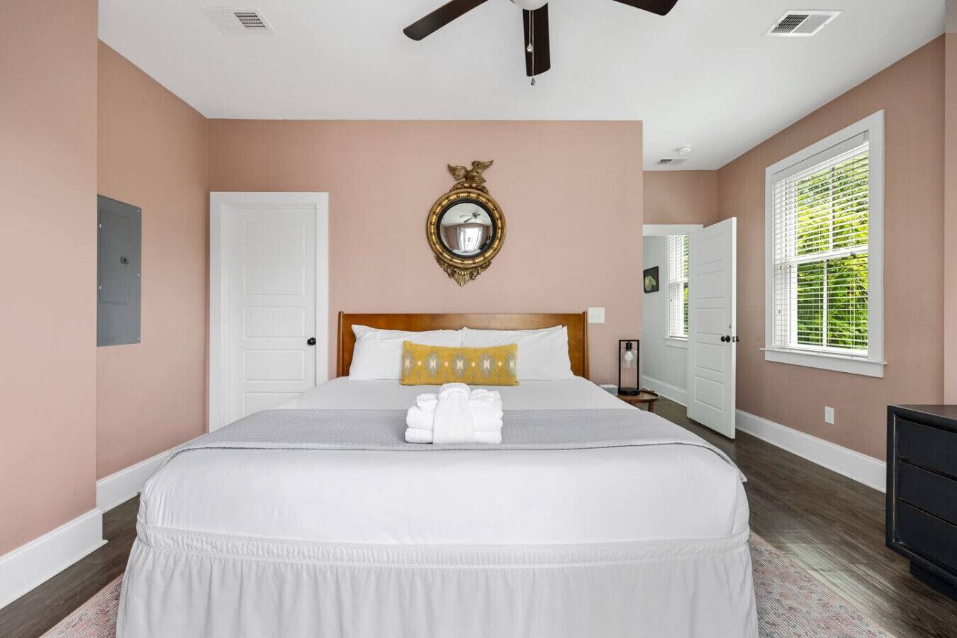 A bedroom with a large bed, a ceiling fan, a mirror, and a window with blinds.