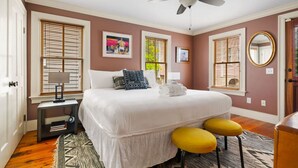 A bedroom with a large bed, two yellow stools, a ceiling fan, and a window with blinds. - 2BR/2BA Downtown Charleston on Spring St + Parking (Charleston)