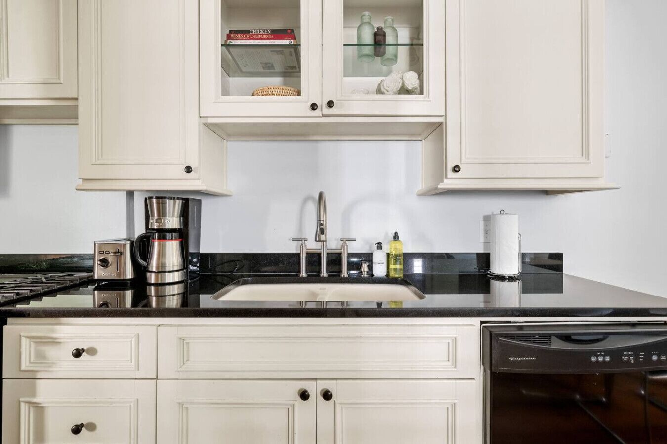 Una cocina con armarios blancos, encimera negra, cafetera, fregadero y lavavajillas.
