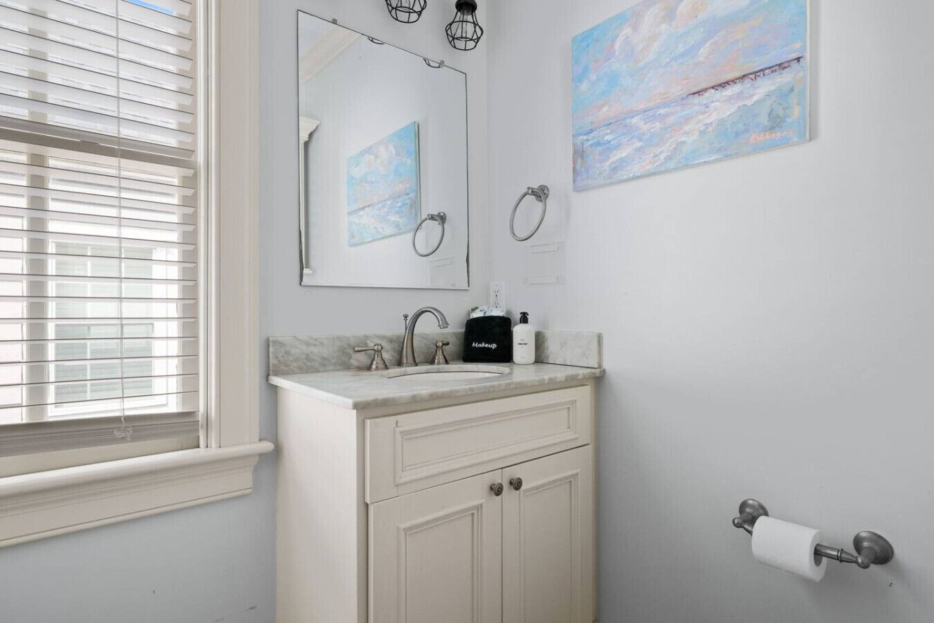 A bathroom with a sink, mirror, towel rings, and a toilet paper holder.