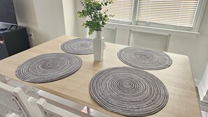 A dining table with four circular placemats, a white vase with a plant, and a window with blinds.