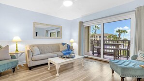 A living room with a sofa, coffee table, and ottoman, a ceiling fan, and a large sliding glass door leading to an outdoor area with a view of palm trees. - Sandy Sea Turtle Pet-Friendly Condo w/ Ocean Views (St. Augustine Beach)