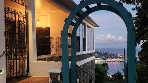 Una casa con una scala in pietra che conduce a un balcone, un arco turchese e vista su uno specchio d'acqua e luci cittadine in lontananza.