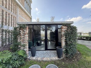 Un área de estar exterior moderna con cerramiento de vidrio, rodeada por un muro de ladrillo y vegetación.