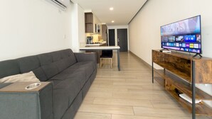 A modern living room with a grey sofa, a wooden TV stand, and a kitchen area in the background. - Dpto. cerca de Ternium y UANL (Monterrey)
