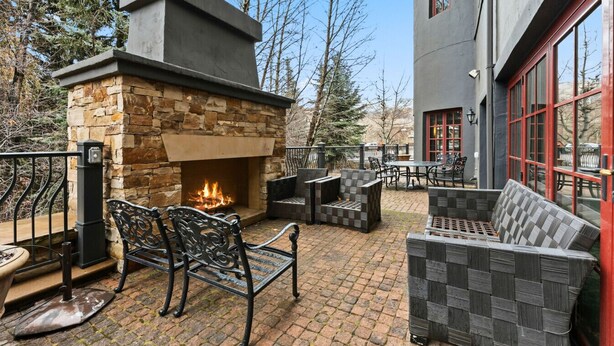 An outdoor patio with a stone fireplace, outdoor furniture, and a view of trees and a building. - Beaver Creek Penthouse | Pool, Hot Tub, Walkable (Avon)