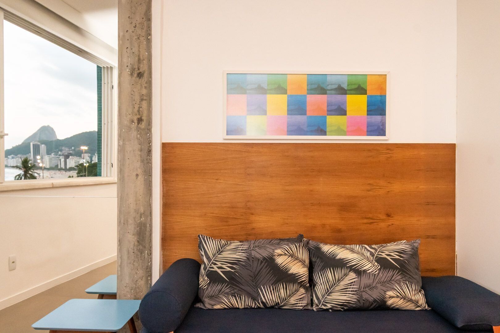 Une chambre avec une tête de lit en bois, un lit agrémenté d’oreillers à motifs, une peinture abstraite aux couleurs vives et une vue sur le paysage urbain par la fenêtre.