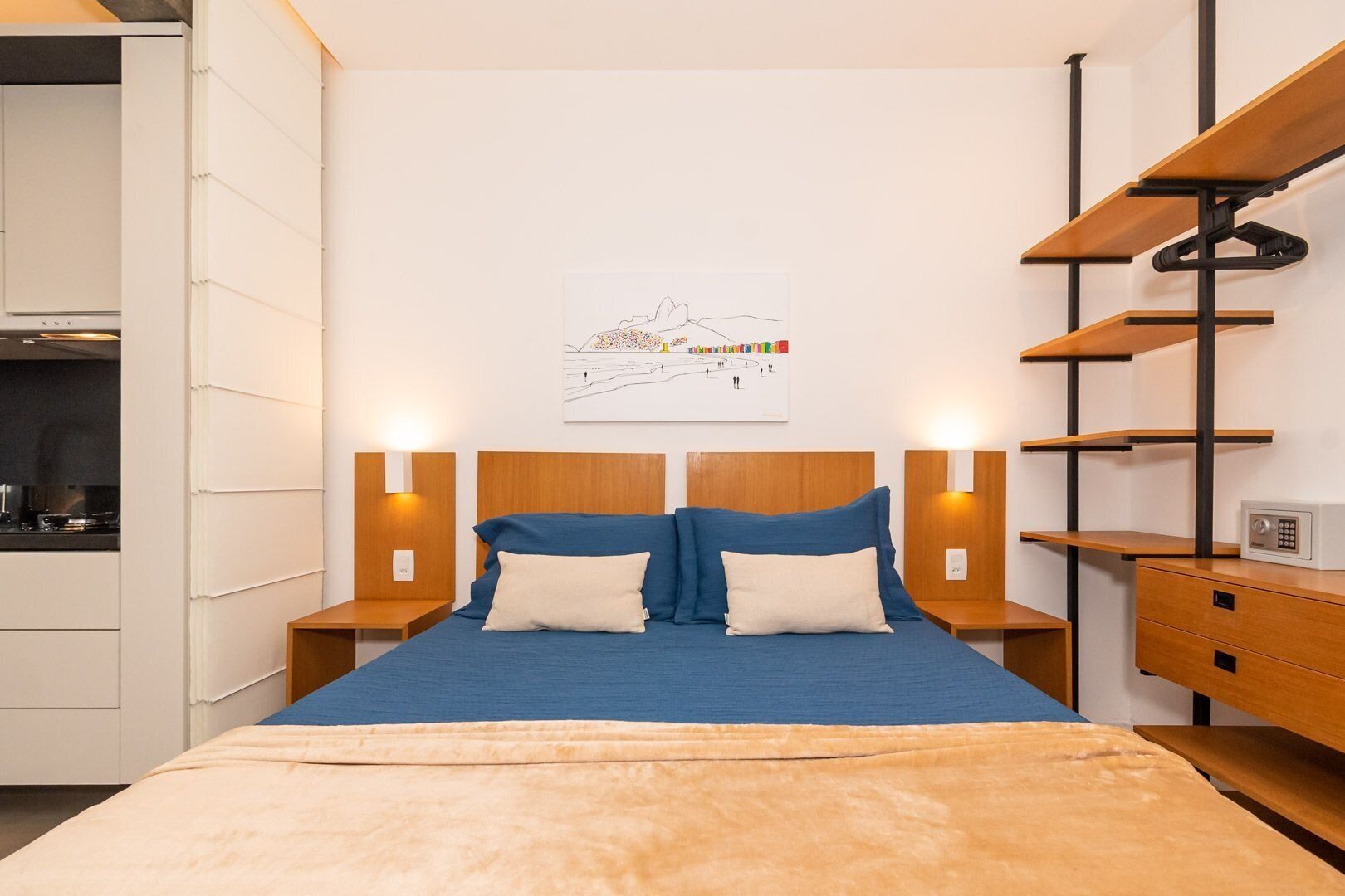 Une chambre moderne avec un lit, des tables de chevet, une étagère fixée au mur et un coin cuisine visible sur la gauche.