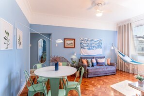 Un salon avec une table ronde, des chaises vertes, un canapé avec des coussins, un hamac et un grand tableau accroché au mur.