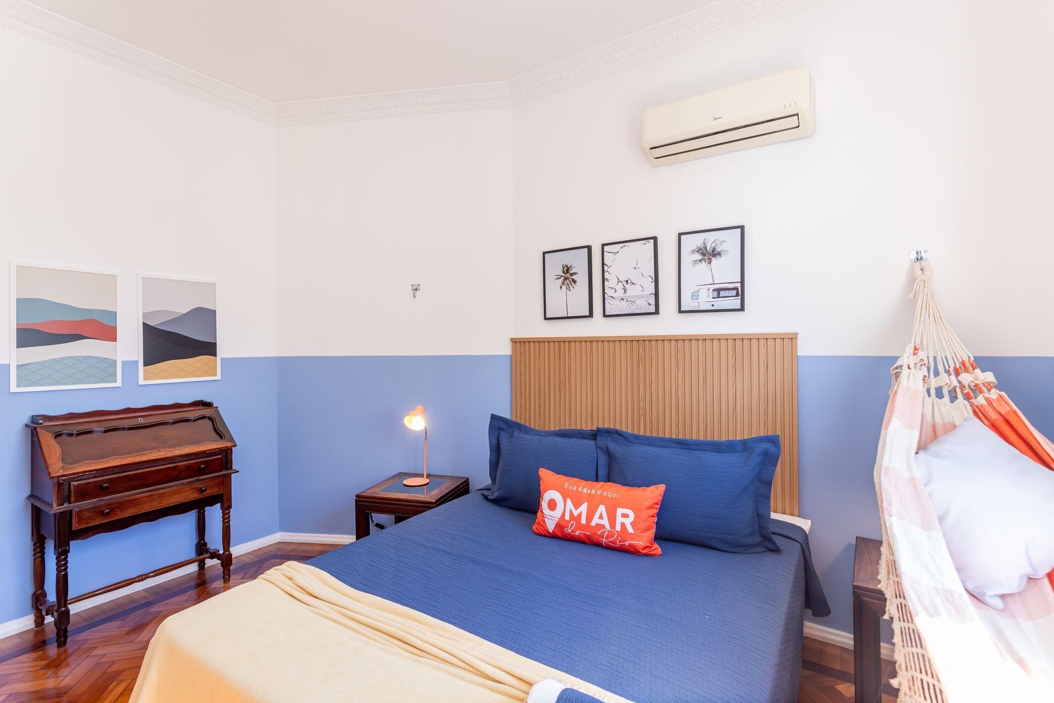 A bedroom with a bed, a wooden nightstand with a lamp, a desk, and wall art.