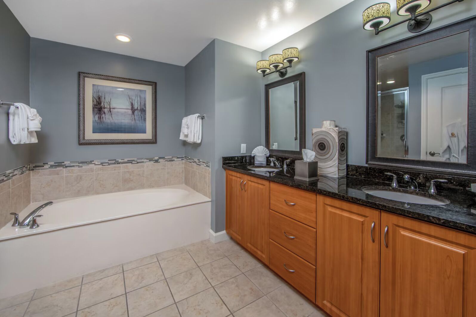 A bathroom with a bathtub, two sinks, wooden cabinets, a framed painting, and a mirror with a light fixture.