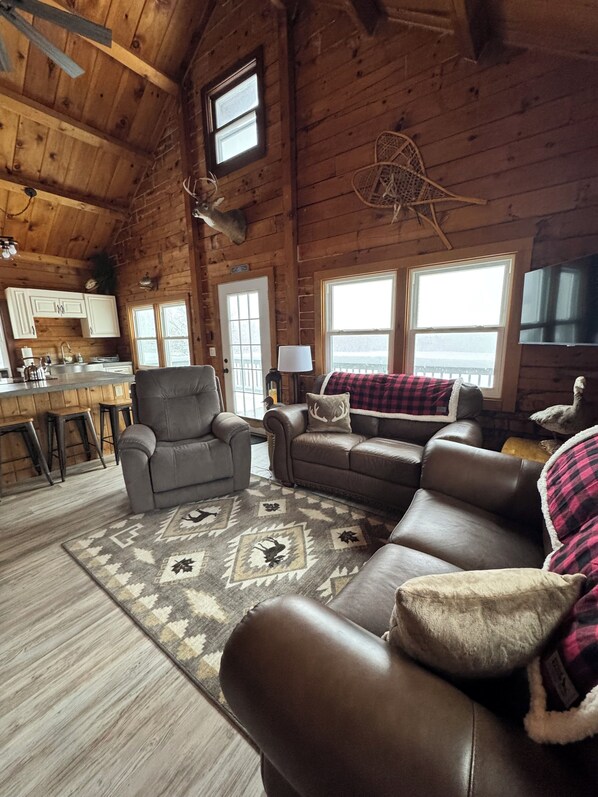 A cozy cabin interior with a wooden ceiling, a kitchen area, a seating area with leather sofas, and a large window providing a view.