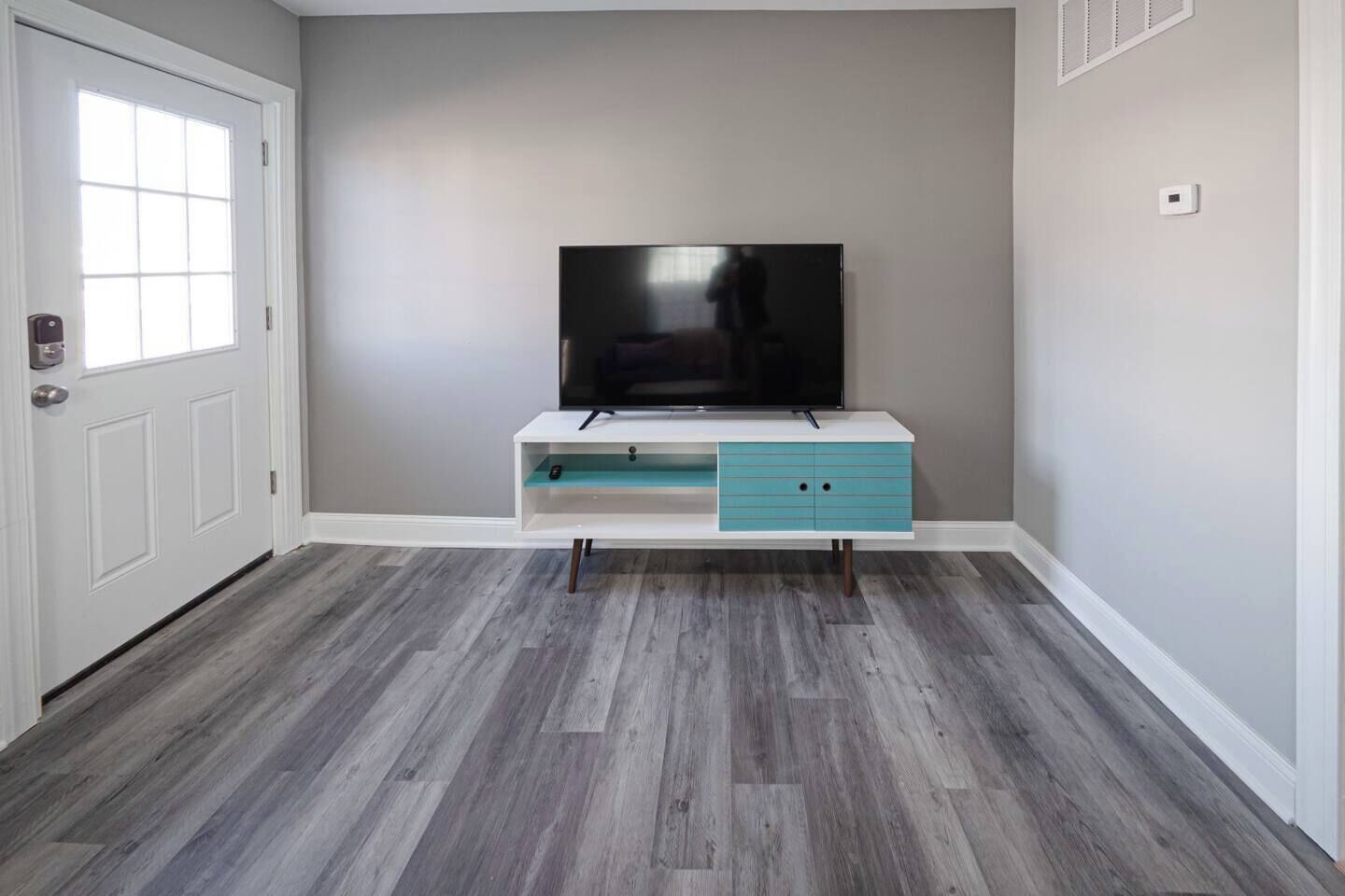 Una habitación con puerta blanca, paredes grises, un televisor de pantalla plana sobre un mueble blanco con cajones color turquesa y suelo de madera gris.