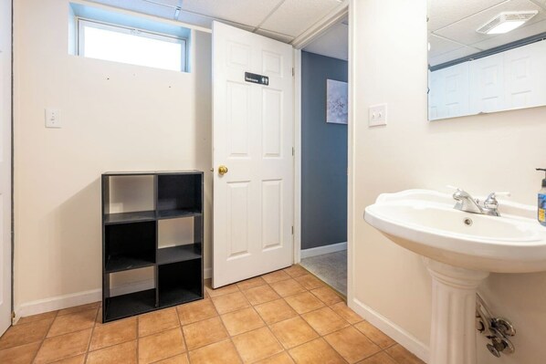 A bathroom with a white sink, a door with a sign, and a small shelf unit. - Charming Modern Farmhouse Retreat (Terre Haute)
