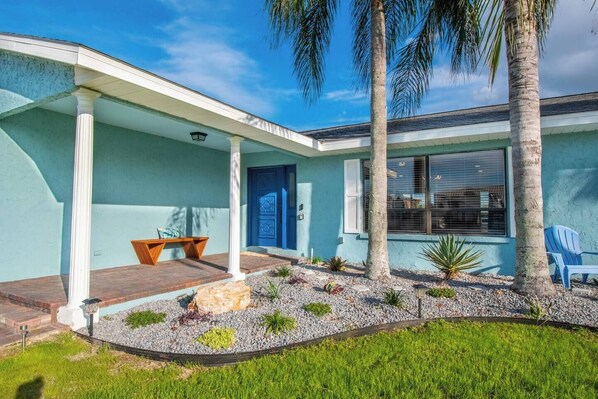 Una casa con porche cubierto, puerta azul y un pequeño jardín con palmeras y un banco.