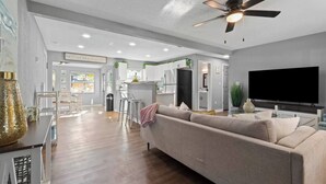Un salon moderne avec un canapé, un téléviseur et un ventilateur de plafond. On aperçoit à l’arrière-plan un coin cuisine avec des meubles blancs et un réfrigérateur.