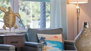 Une pièce avec vue sur la verdure, un fauteuil gris agrémenté d’un coussin aux motifs tropicaux, une petite table et une figurine de tortue décorative posée sur une table de chevet.
