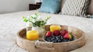 Cama bem arrumada com uma bandeja de madeira com frutas frescas e suco, mesa de cabeceira com abajur, e um painel decorativo na parede.