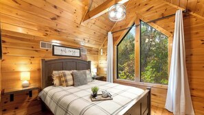 Une chambre confortable aménagée dans un chalet en bois, avec un grand lit, une table de chevet avec une lampe, une fenêtre avec des rideaux et un panneau décoratif au mur.
