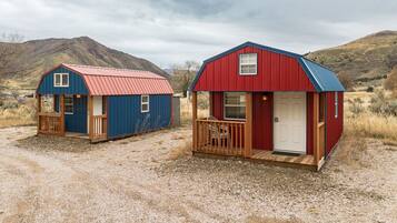 Deux petites cabanes aux façades rouge et bleu, dotées de terrasses en bois et entourées d’un sol en gravier.