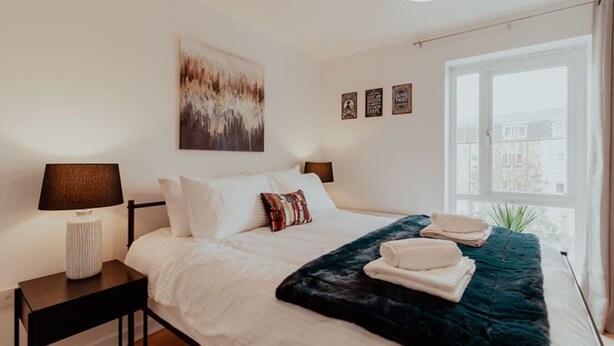 A neatly arranged bedroom with a bed, two bedside tables, a window with a view, and a wall-mounted artwork. - The Dickens Den | 1 Bed Flat in Rochester for 4 (Medway)