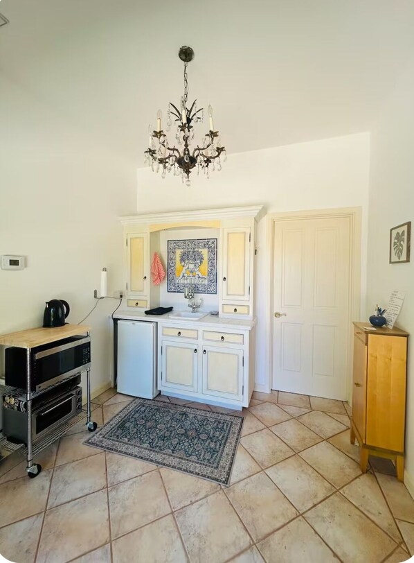 A compact kitchen with a microwave, kettle, and a small refrigerator. A chandelier hangs from the ceiling, and a patterned rug is on the floor.
