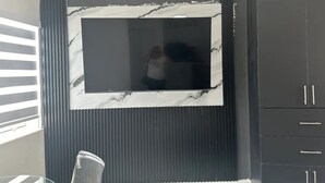 Ein Zimmer mit einem großen Flachbildfernseher an einer Wand mit Marmoroptik, einem dunklen Schrank zur Rechten und einem Fenster mit Jalousien zur Linken.