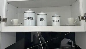 Ein weißes Schränkchen mit drei beschrifteten Behältern: Kaffee, Tee und Zucker.