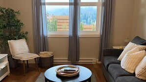 Ein Wohnzimmer mit rundem Couchtisch, einer Couch, einem Sessel, einer Pflanze und Blick ins Freie durch große Fenster.