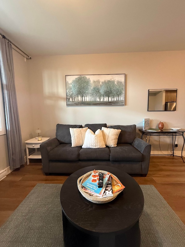 Un salon moderne avec un canapé gris foncé, une table basse ronde sur laquelle sont posés des magazines, un tableau représentant des arbres, une petite table d’appoint et un miroir au mur.