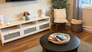 Ein modernes Wohnzimmer mit einem an der Wand befestigten Flachbildfernseher, einer weißen Unterhaltungsecke, einem runden Couchtisch mit Zeitschriften, einer Topfpflanze und einem Fenster mit Vorhängen.
