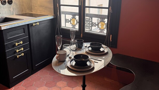 Un'area pranzo con un tavolo apparecchiato per due, una finestra con vista sugli edifici e una cucina con pensili neri e pavimento piastrellato.