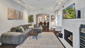 A spacious living room with a gray sectional sofa, a coffee table, and a fireplace. There is a dining area in the background with a table and chairs. - Private Tropical Garden Home - Walk to Las Olas (Fort Lauderdale)