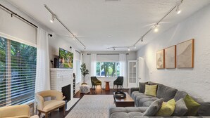 A spacious living room with a fireplace, a grey sofa, and a dining area with two chairs. - Private Tropical Garden Home - Walk to Las Olas (Fort Lauderdale)